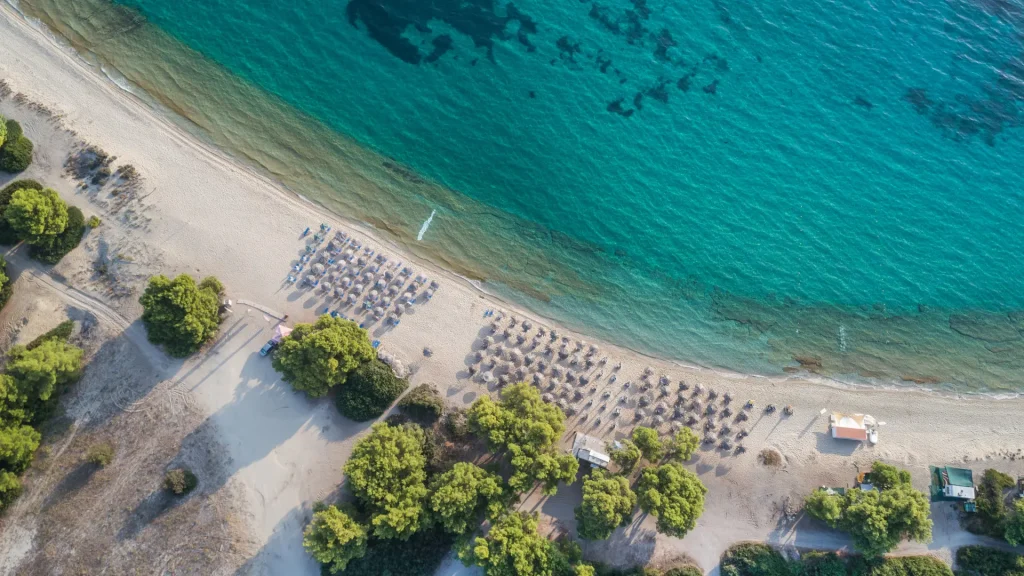  Aerial view of a Naxos beach lined with sun loungers and umbrellas, bordered by turquoise Aegean waters and shaded by tamarisk trees.