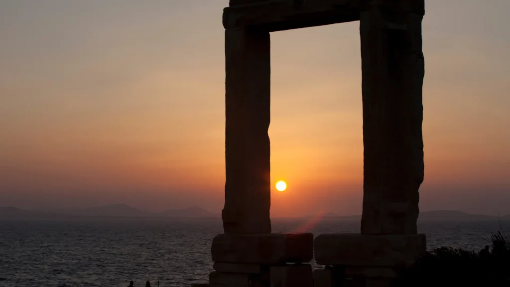 Silhouetted Portara marble gateway framing a golden sunset over the Aegean Sea.